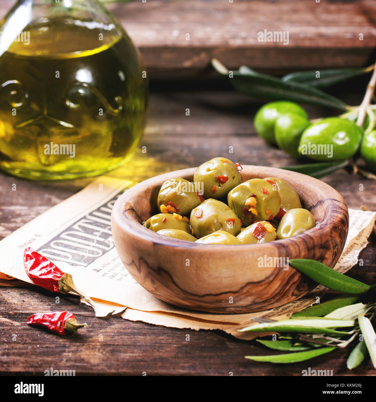 Green olives in olive wood bowl and bottle of olive oil served with chili peppers on old wooden table. Square image with selective focus Stock Photo