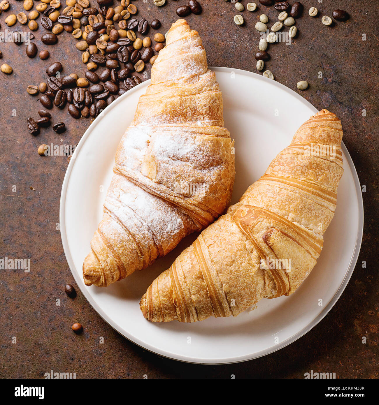 Plate with two fresh baked croissants with heap od roasted and  unroasted coffee beans over rusty metal background. Square image Stock Photo