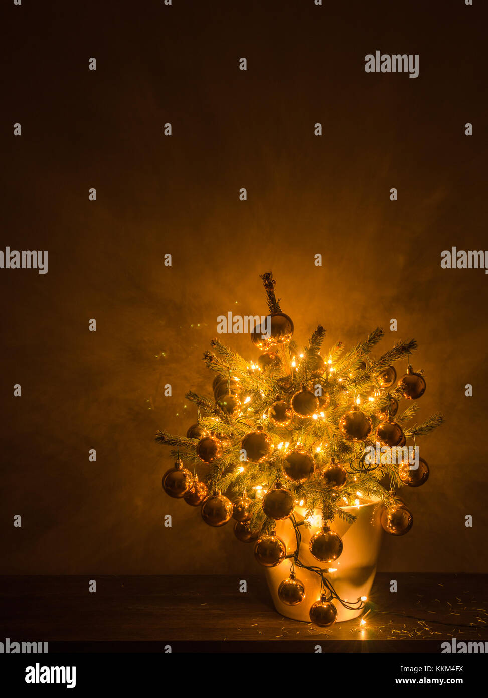 Small Christmas tree with luxury baubles and warm lights, in a white plastic flowerpot, with the dark background. Stock Photo