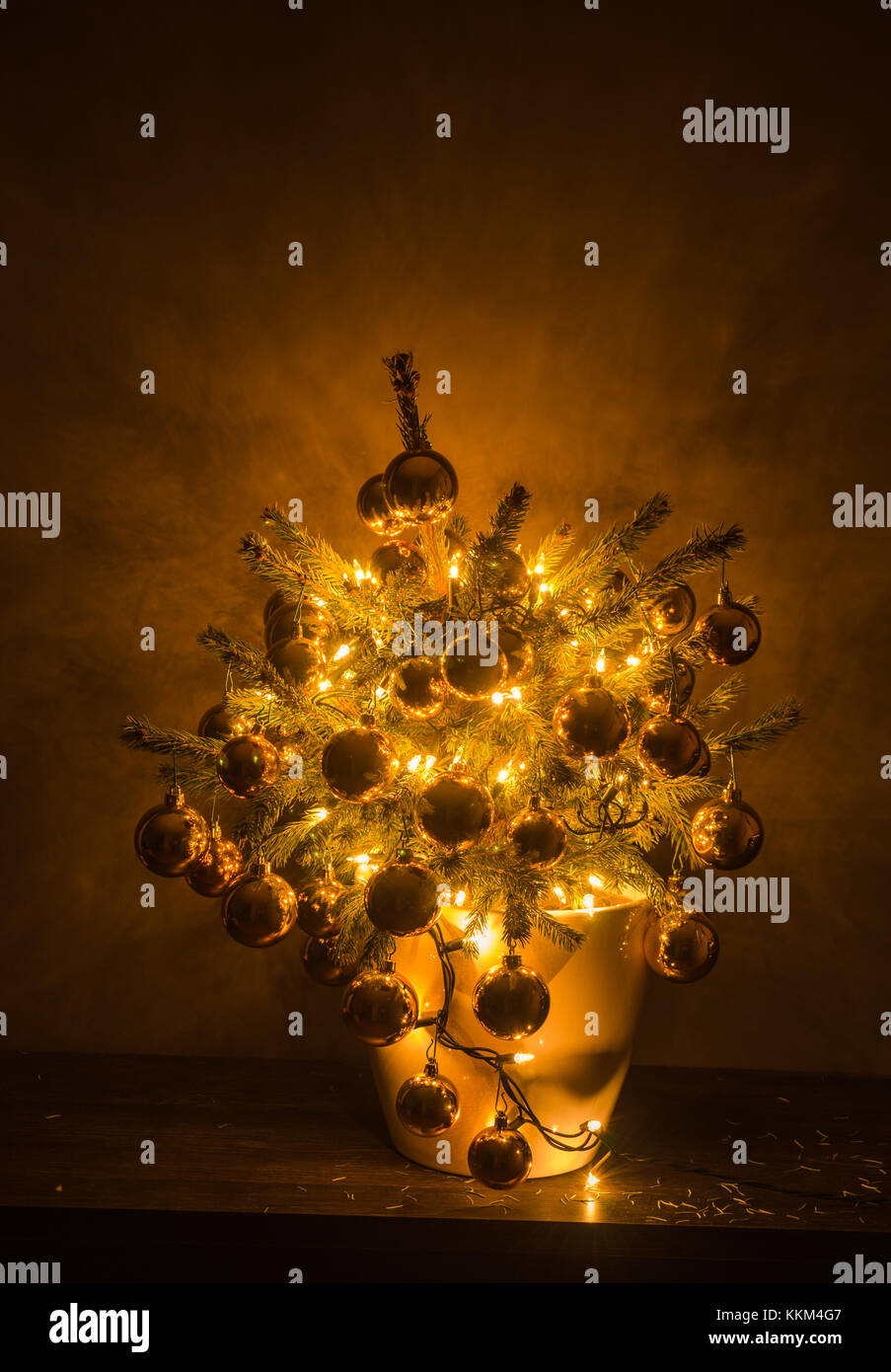 Small Christmas tree with luxury baubles and warm lights, in a white plastic flowerpot, with the dark background. Stock Photo