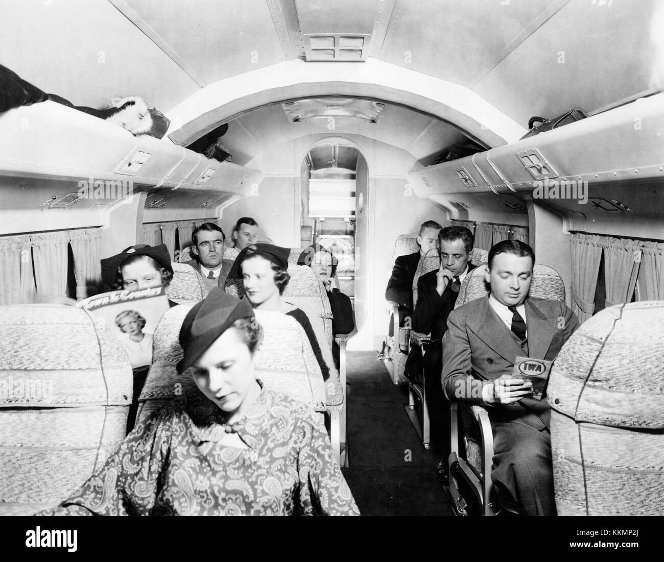 Posed interior view of passenger cabin of a Pan American Airways (PAA) Sikorsky S-43A Amphibion (Baby Clipper) with seated passengers. Rear access door is open showing a Pan American Airways life ring stowed behind the ladder. Note man at right foreground reading a TWA timetable. Circa 1936. Interior view of a Pan Am Sikorsky S-43A (Baby Clipper) Stock Photo