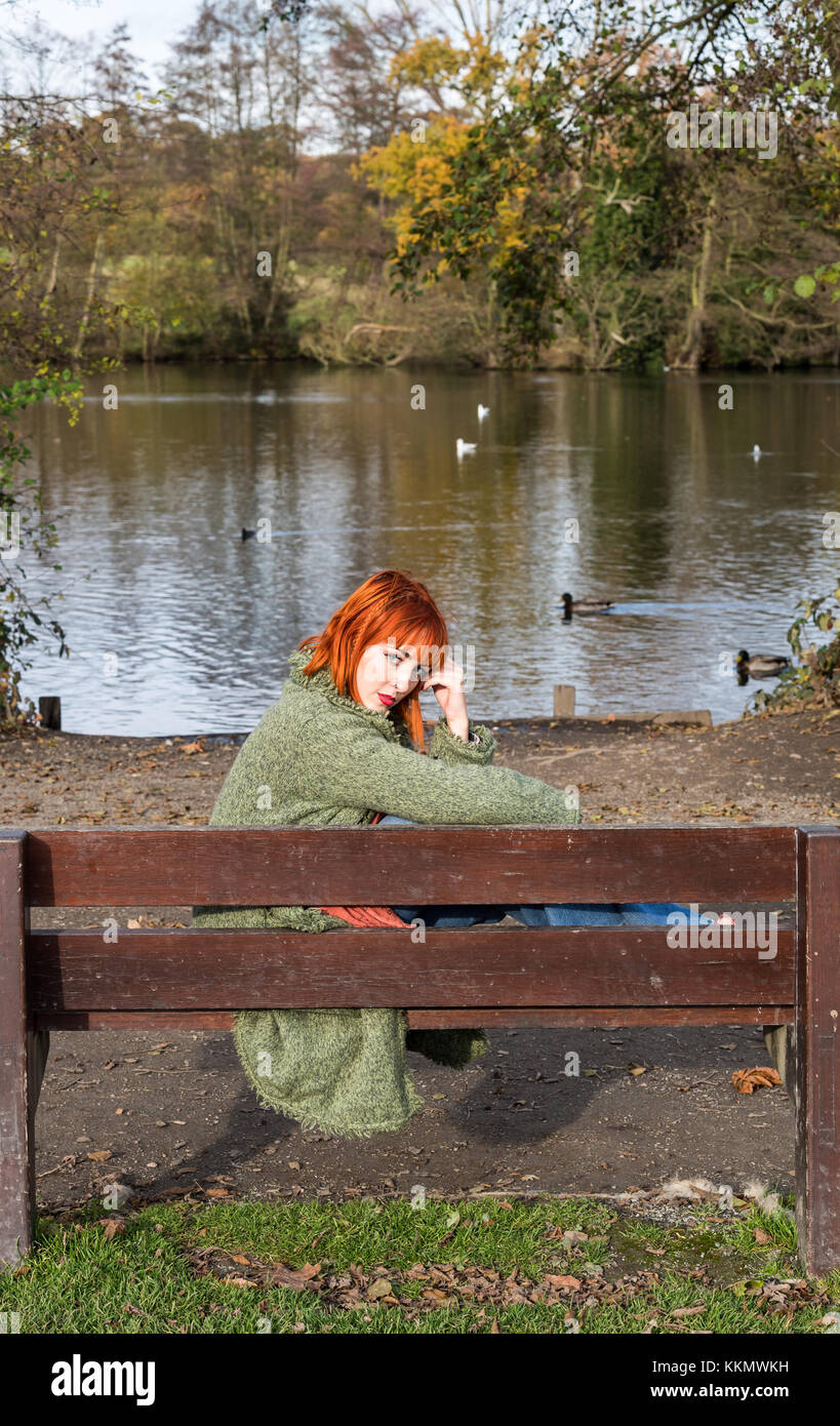 Redhead Girl on bench in park beside lake  in autumn Stock Photo
