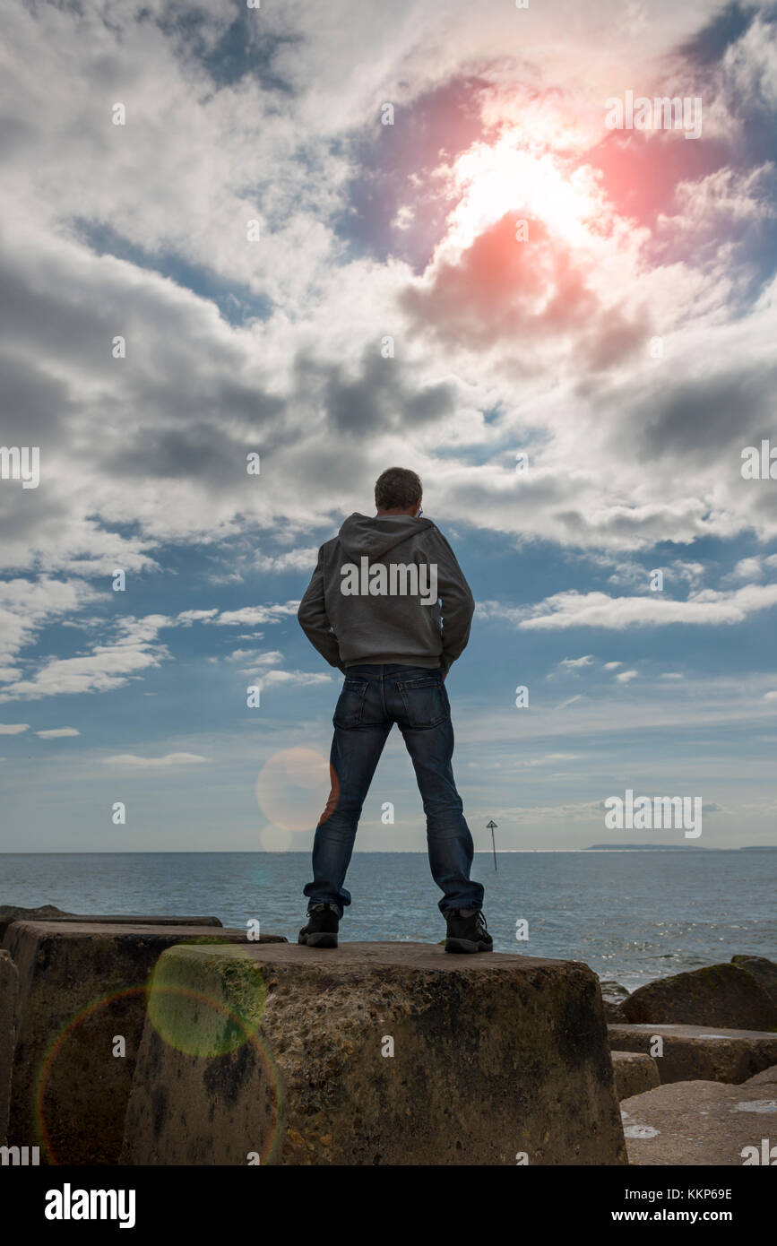 back view of a man standing on sea defence looking out to sea. Lens flare. Stock Photo