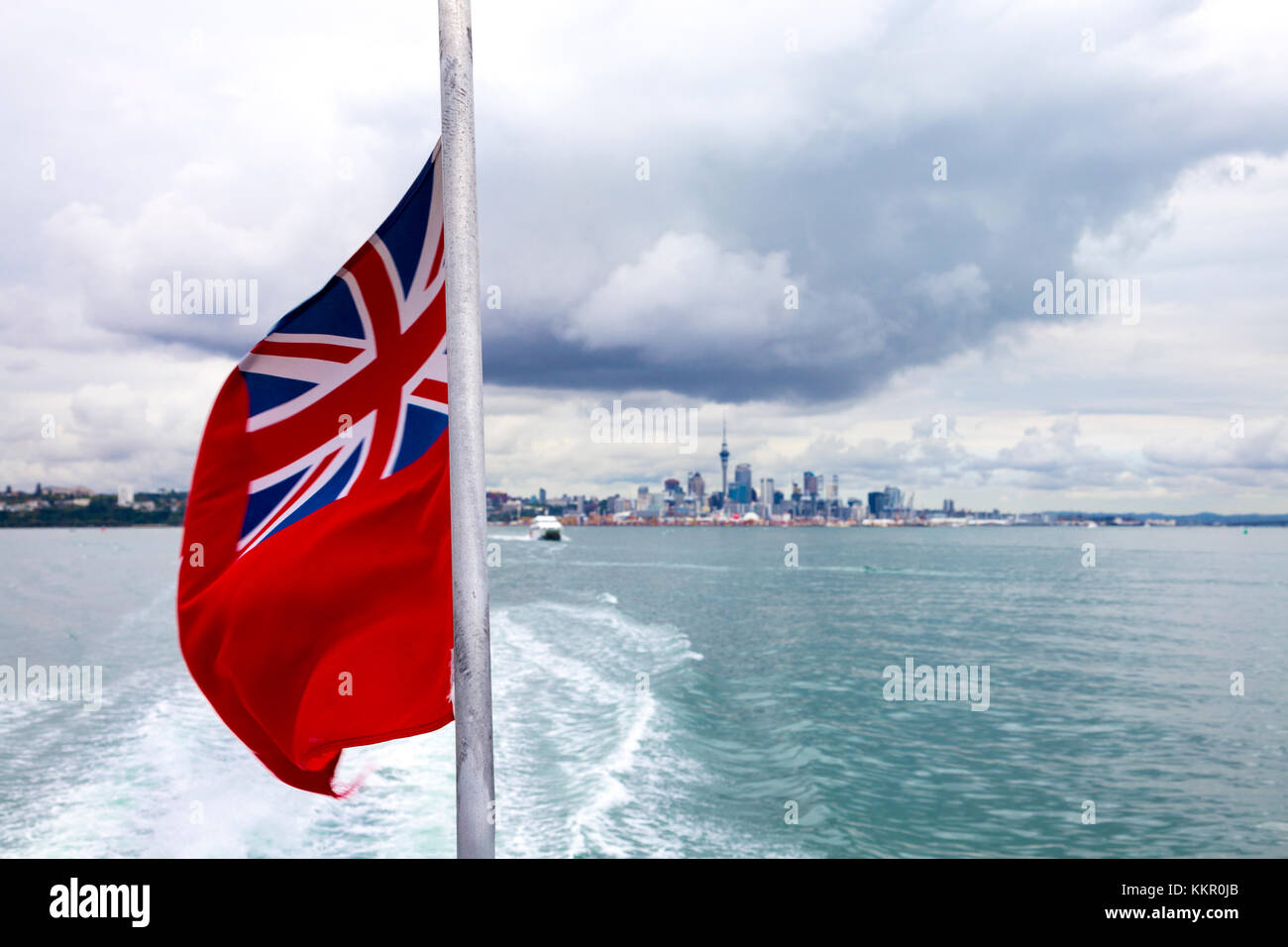 Red ensign flag at the stern of a boat waving in the wind with skyline of Auckland in the background, New Zealand Stock Photo