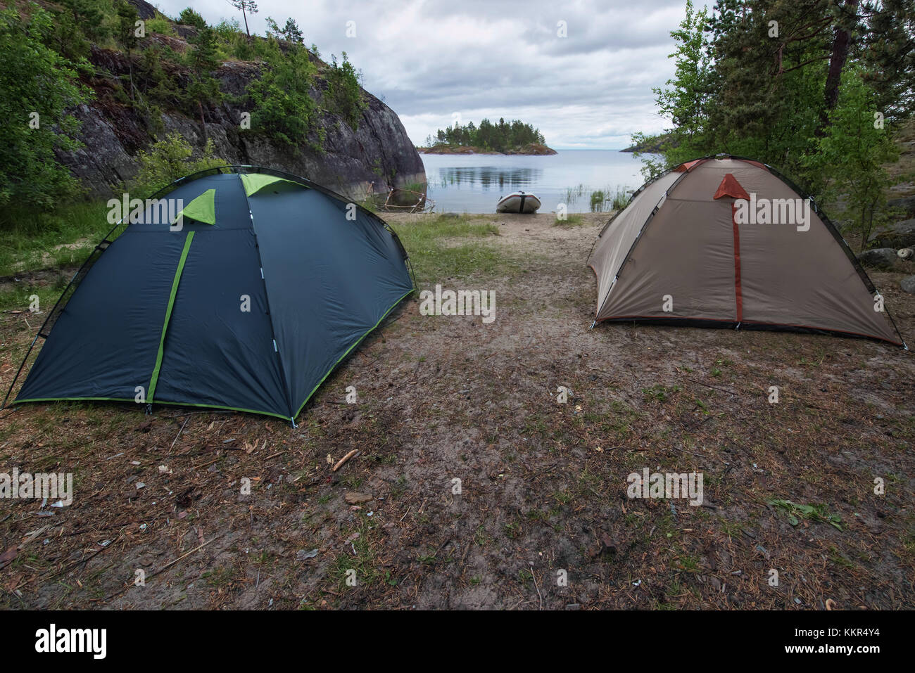Tourist camp with two tents and inflatable boat at the river or lake bank. Camp is located among the forest and rocks. Summer cloudy weather. Stock Photo