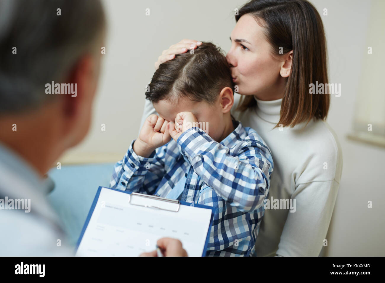 Young woman kissing her little crying son during visit to doctor Stock Photo