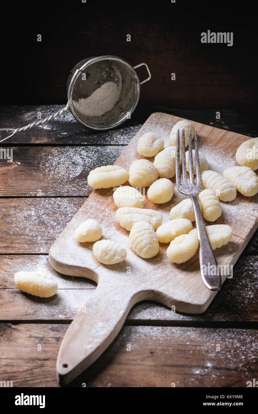 Uncooked homemade potato gnocchi with fork and strainer on vintage cutting board over wooden table with flour. See series Stock Photo