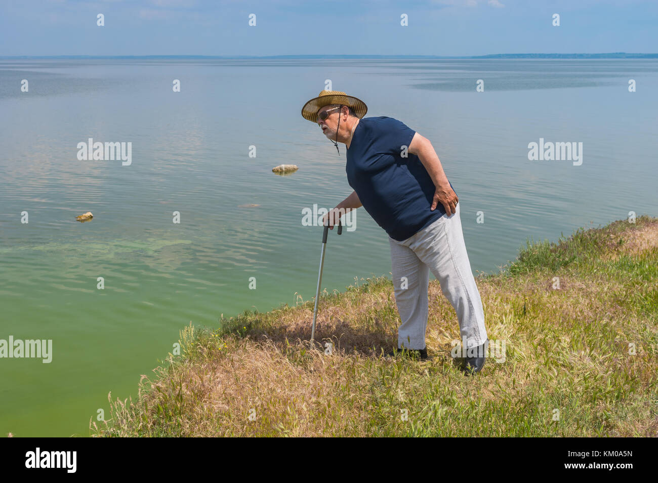 Lonely senior man in blue t-shirt and light pants standing on elevated abrupt riverside of the Dnipro river and looking down Stock Photo