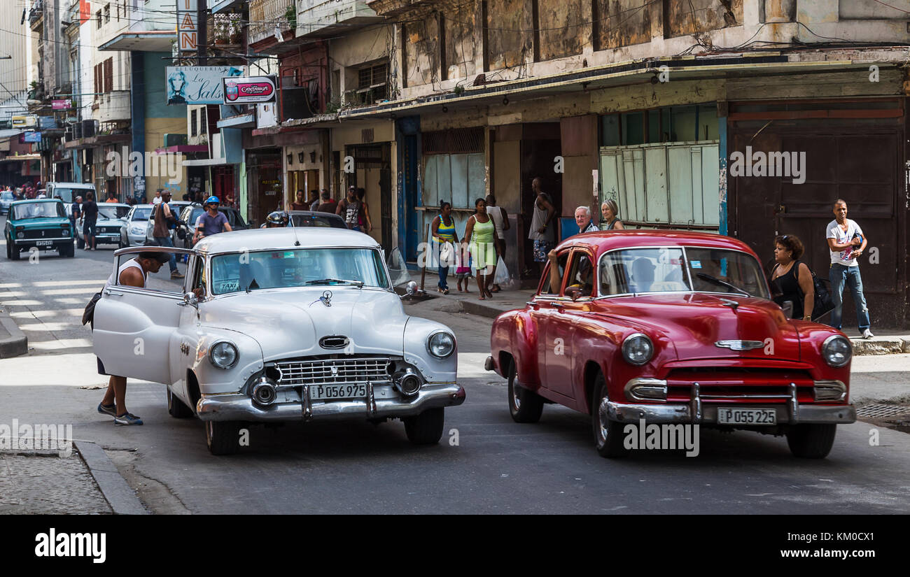 A Cuban man collects his belongings from the back seat of a white classical car as he exits in Centro Havana.  The background is typical busy street i Stock Photo
