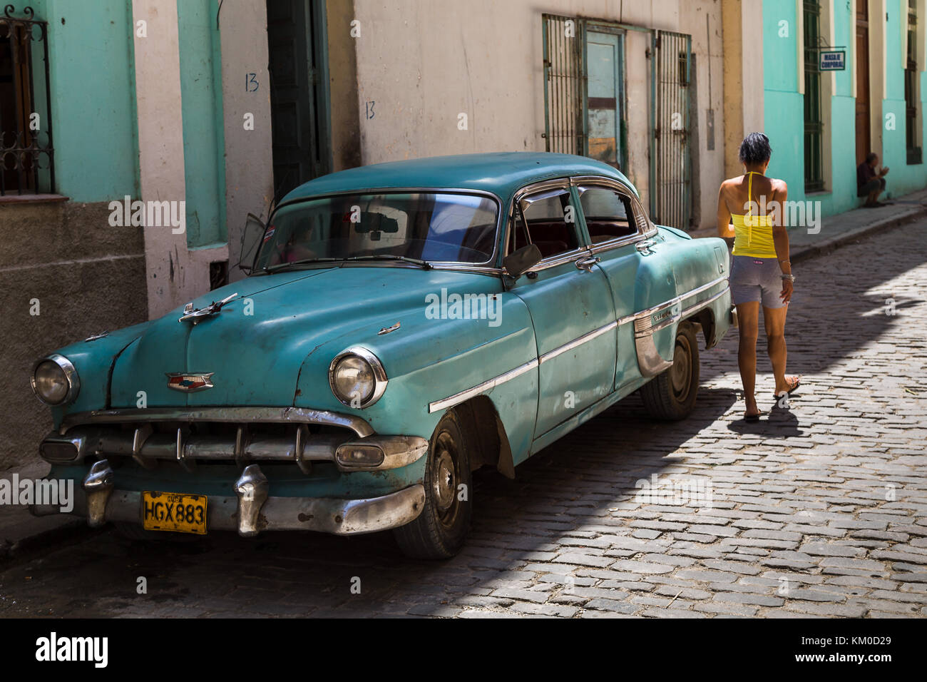 A Cuban woman in colourful clothing walking down a typical backstreet in Havana including an old car and pastel coloured buildings.  Captured during A Stock Photo