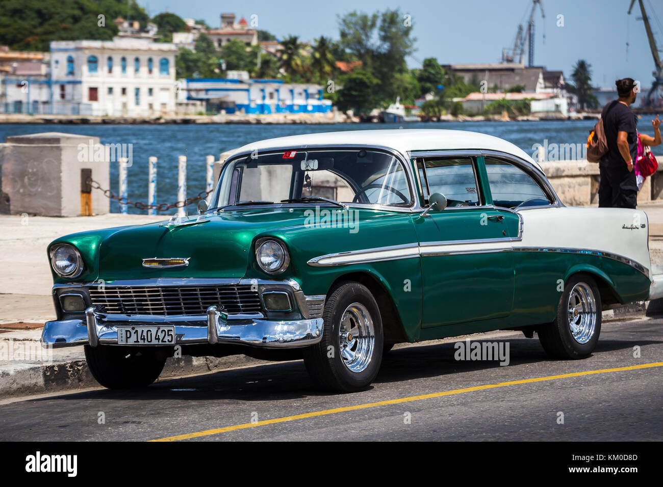 An old American car in prestine condition sits on the side of the road one afternoon in Havana.  The industrial side of the city and harbour can be se Stock Photo