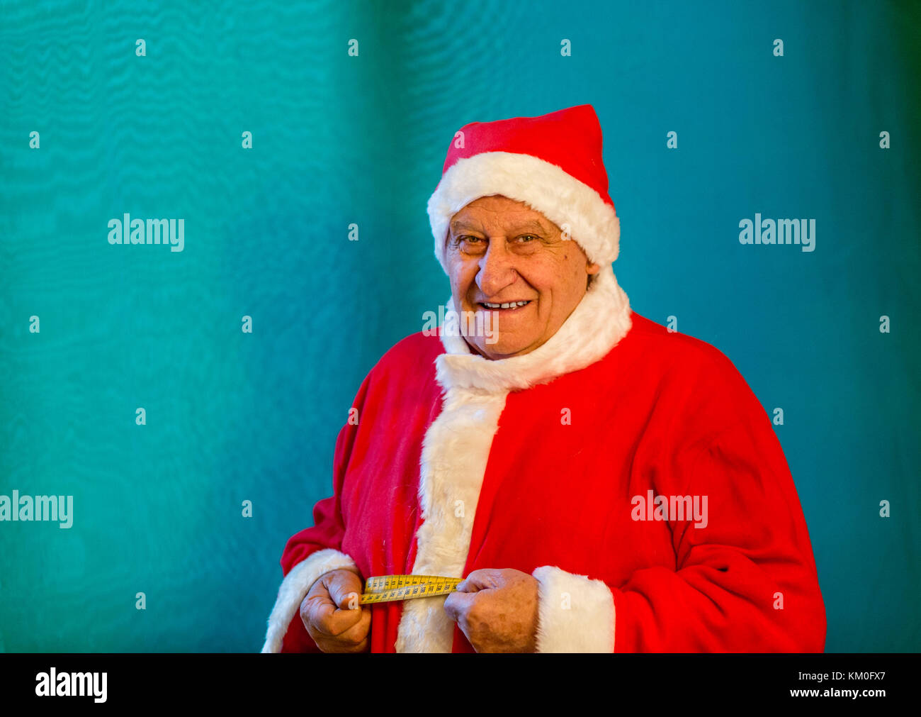 old man wearing Santa Claus costume smiling and measuring the circumference of his belly with a tape measure Stock Photo