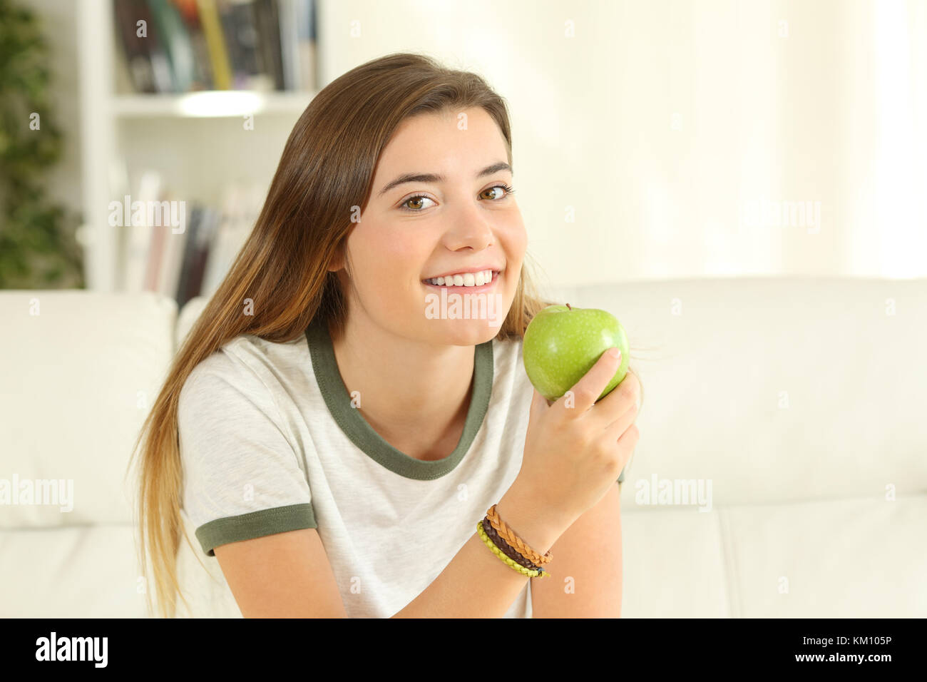 Portrait of a happy teen looking at you holding an apple sitting on a sofa in the living room in a house interior Stock Photo