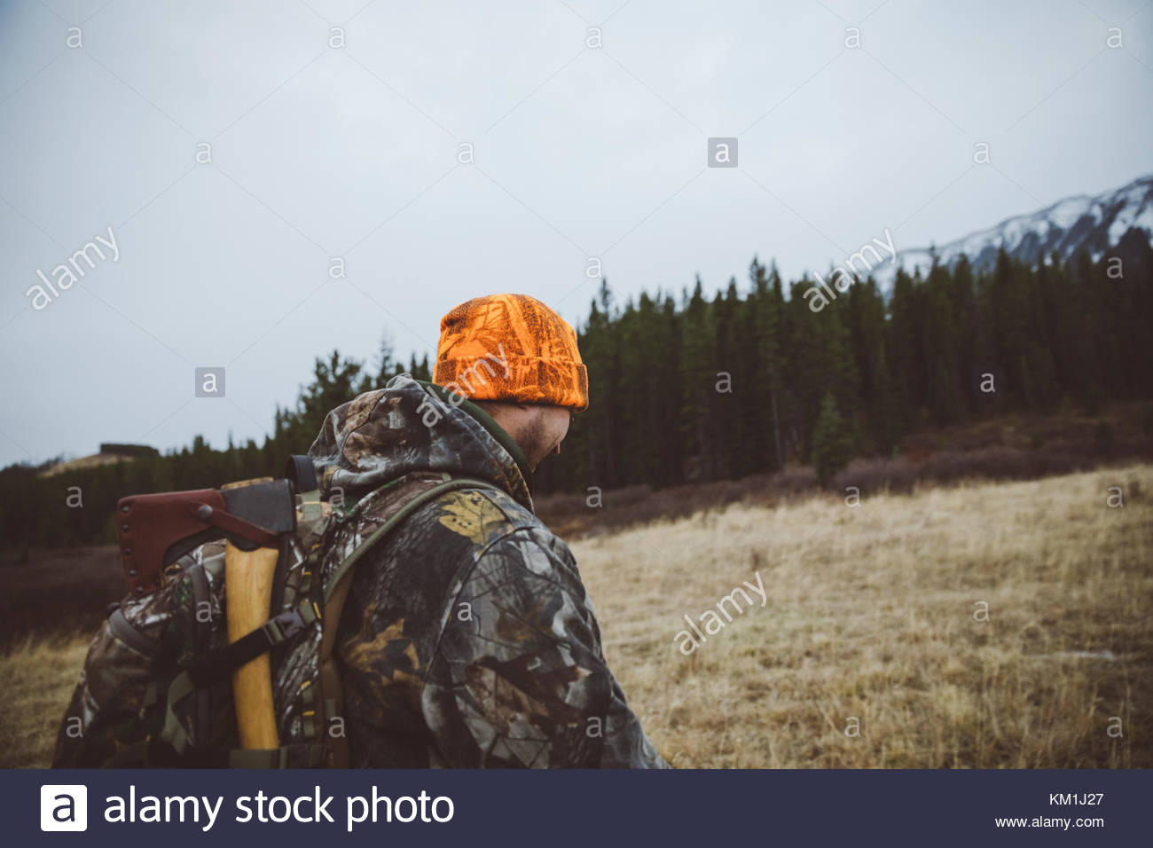 Male hunter in camouflage and orange beanie with hunting backpack and ax in remote field Stock Photo