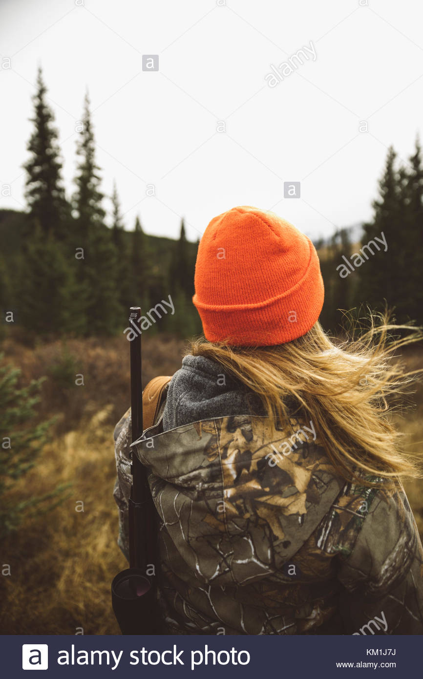 Female hunter in camouflage and orange beanie carrying hunting rifle in field Stock Photo