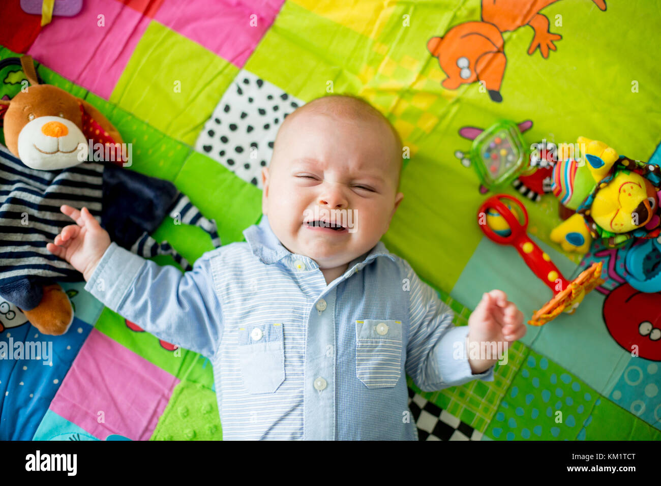 Three months old baby boy, crying at home on a colorful activity blanket, toys and different activity around him Stock Photo