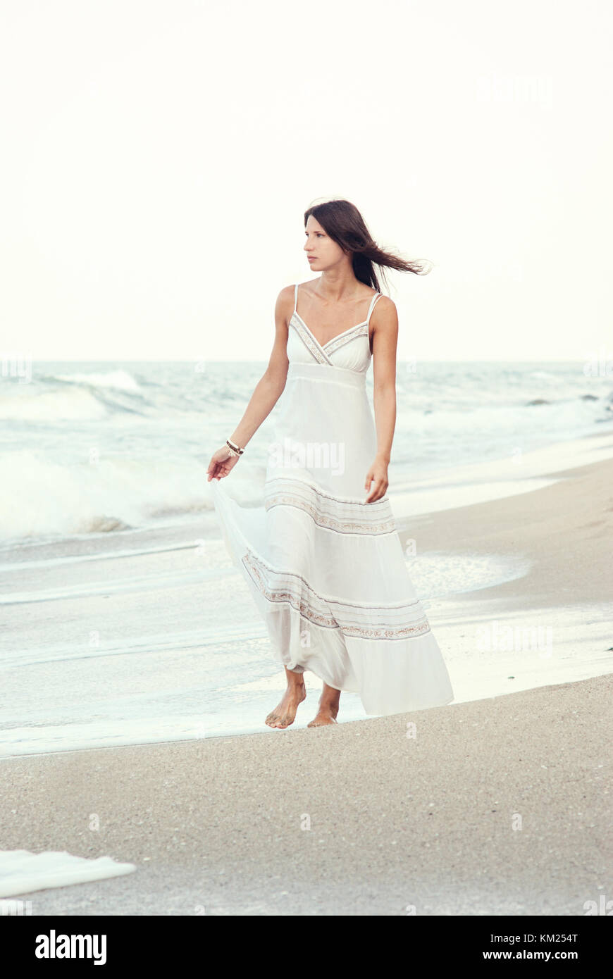 Young beautiful woman in white dress walking on the beach, looking at ...