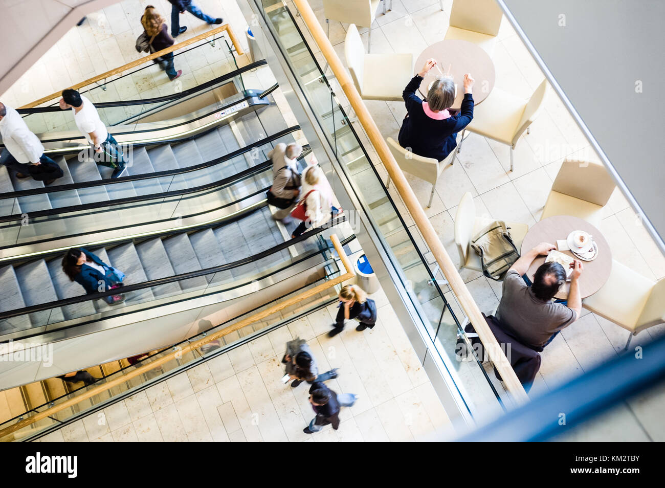 View from above of people in a shopping center taking the escalators, strolling along the walkways and having a snack on the patio of a cafe. Stock Photo