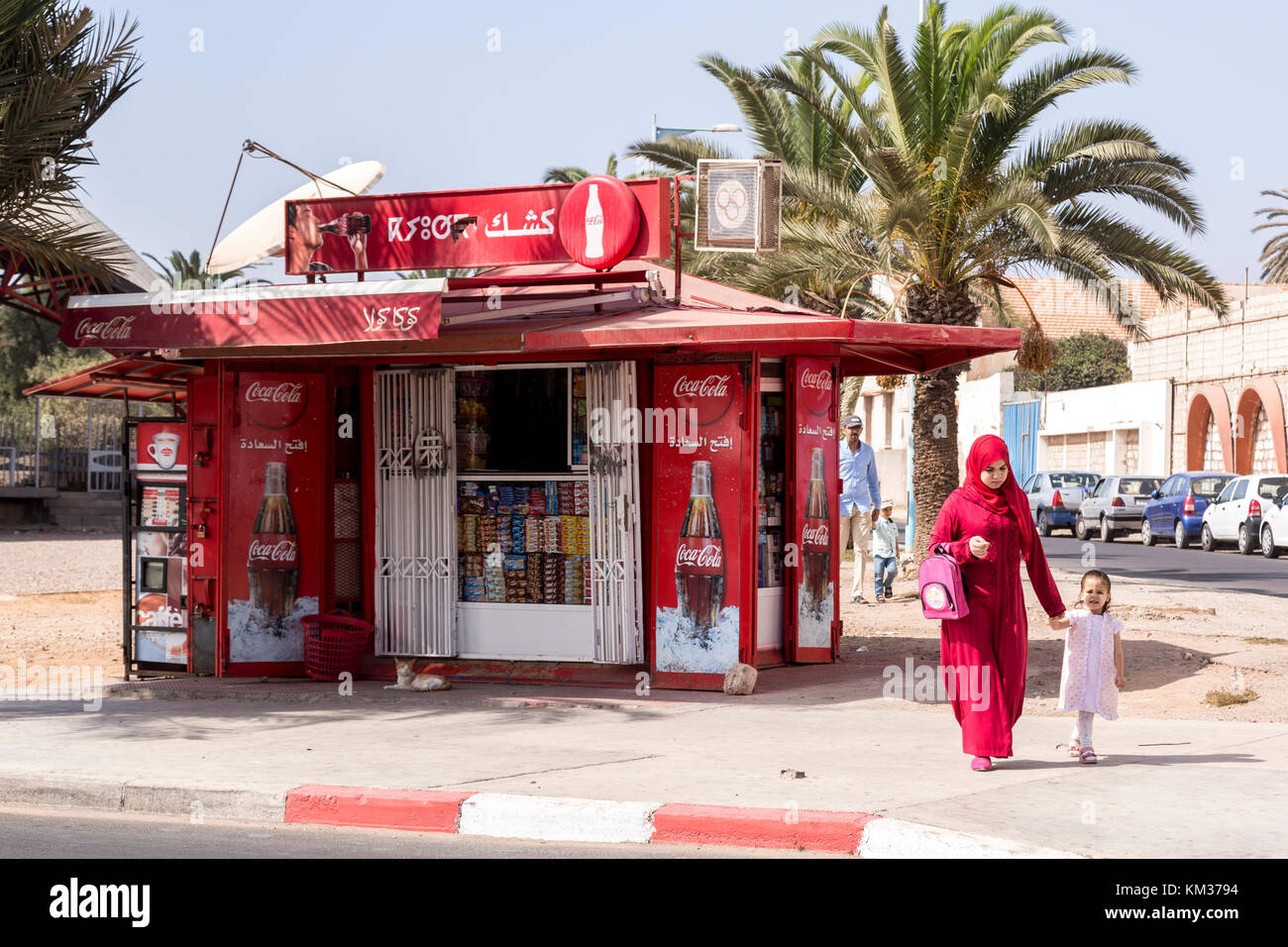 Agadir, Morocco, October 23, 2017: Muslim mother in red hijab walks past red Coca Cola kiosk. Stock Photo