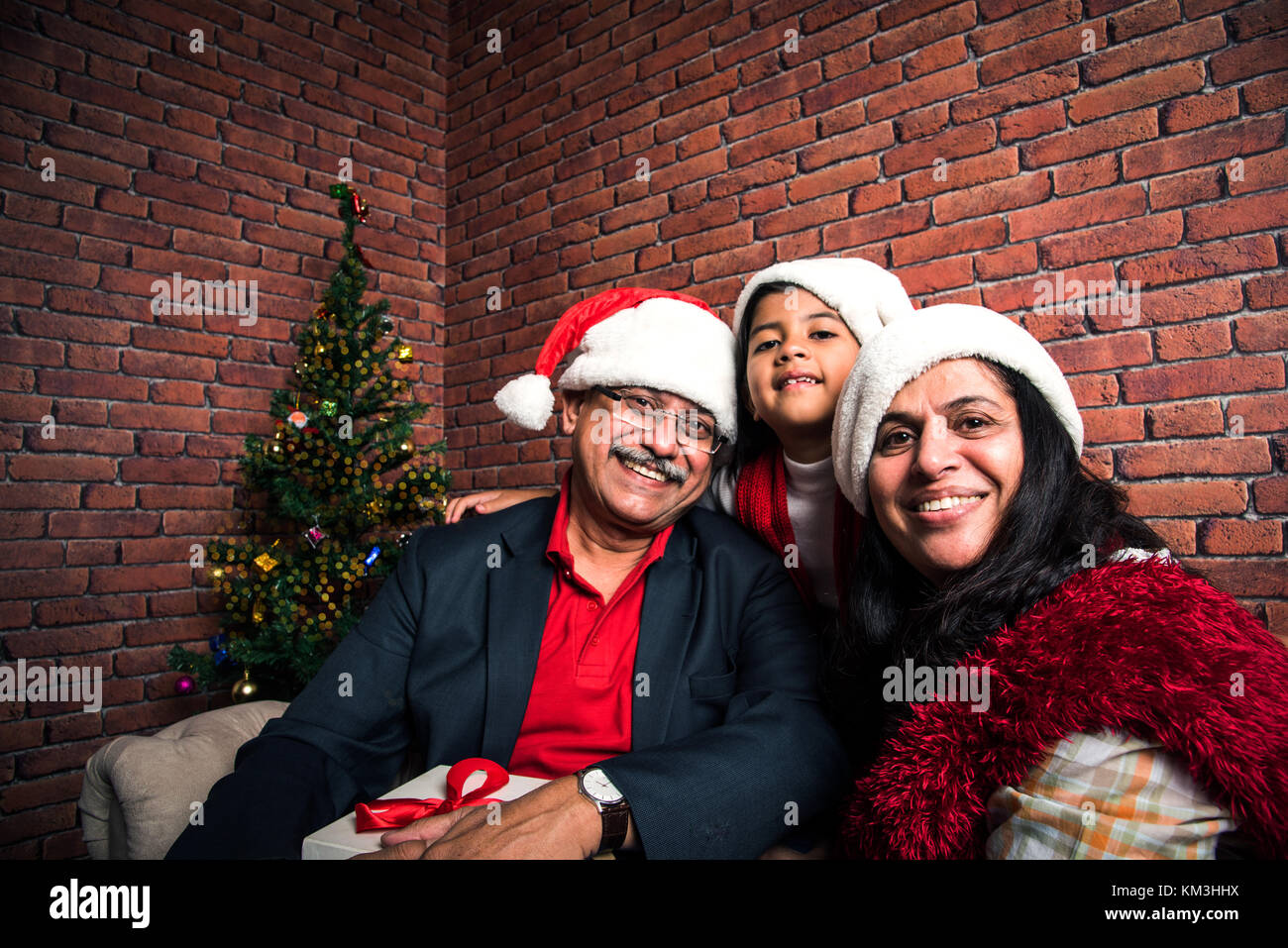 family, holidays, generation, christmas and people concept - smiling Indian grandparents and granddaughter with gift boxes sitting on couch at home Stock Photo