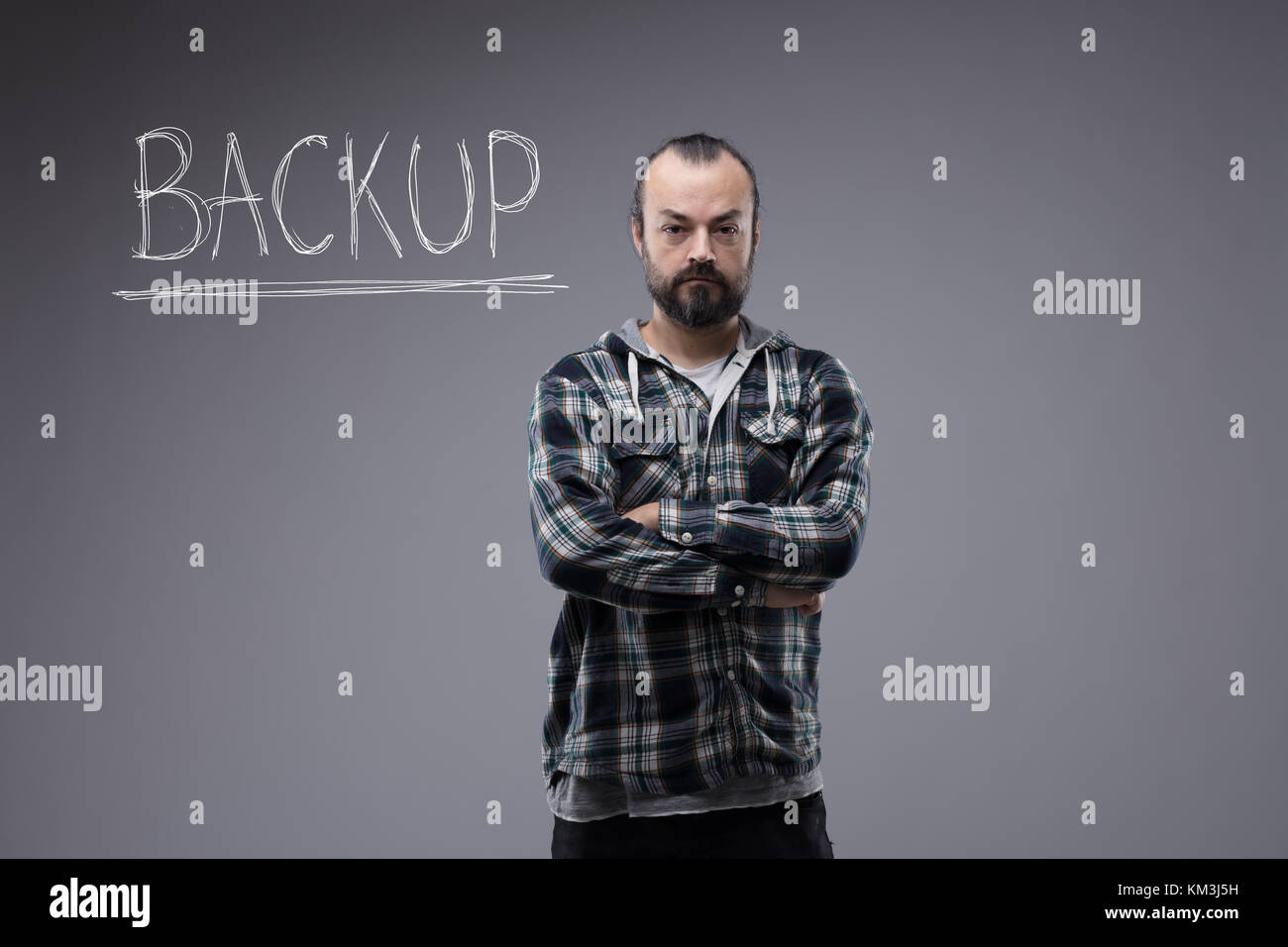 Confident bearded man in a plaid shirt standing with folded arms and a sign Backup handwritten on a blackboard behind him with copy space below Stock Photo