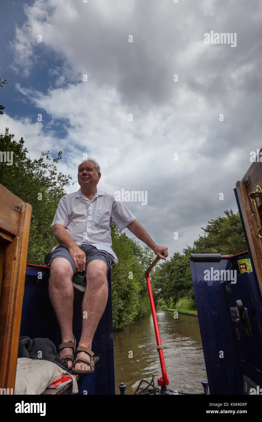 A man in shorts and short-sleeved shirt sits at the rear of a narrowboat whilst steering with the tiller, Coventry canal, Staffordshire, England Stock Photo