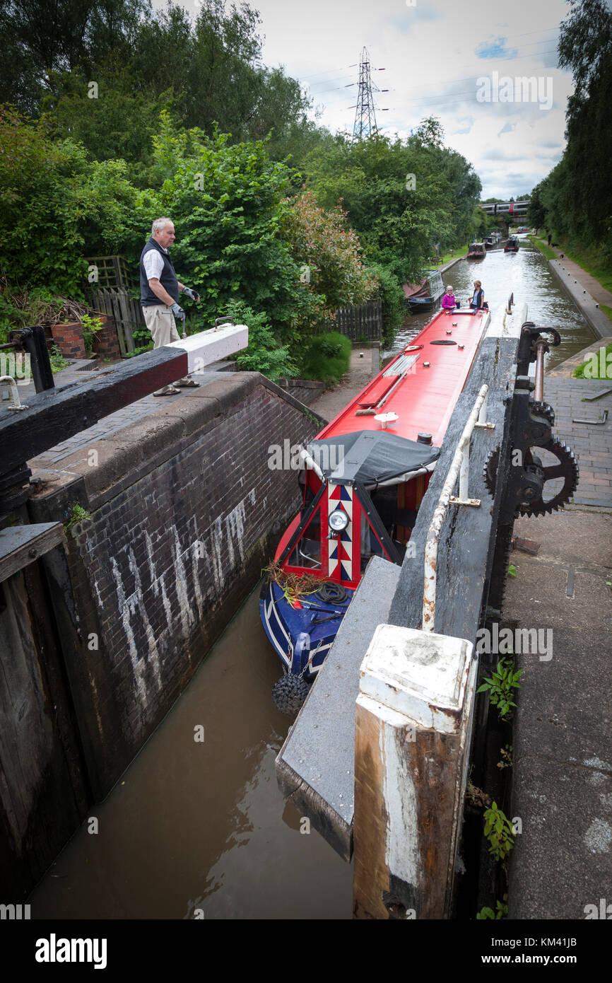 Two women steer their  narrowboat into a lock whilst a man prepares to operate the lock mechanism on the Coventry canal, Staffordshire, England. Stock Photo