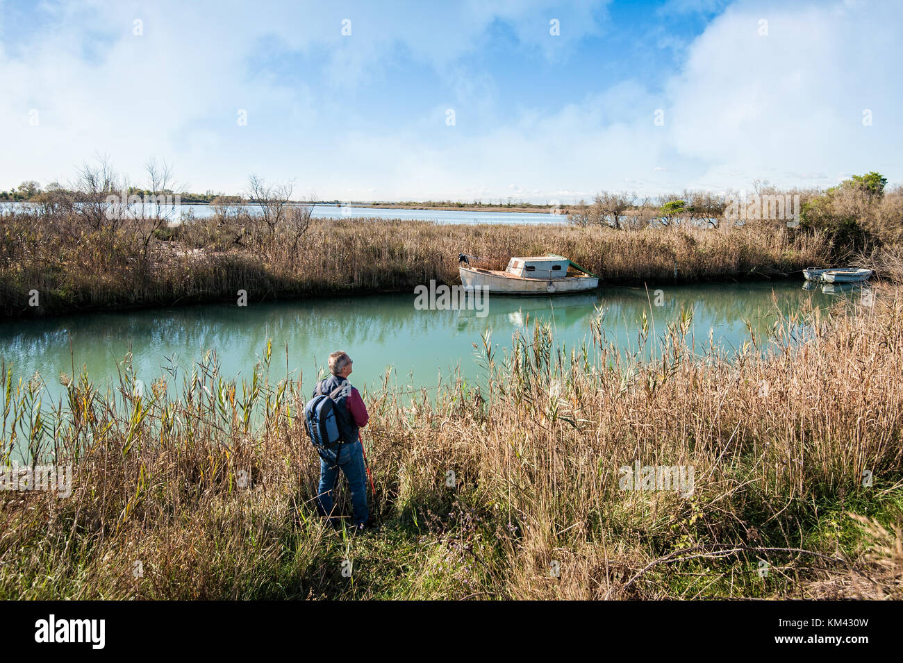 Hiker (about 60 years old) in the wild area of a lagoon. Fishing boats moored along a canal in the lagoon. Stock Photo