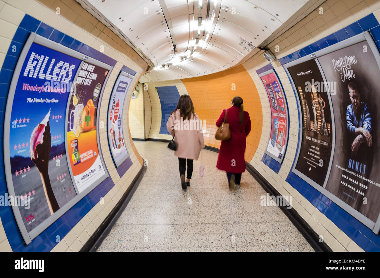Two ladies walk through a connecting tunnel on the London Underground. The walls are covered with advertising posters. Stock Photo