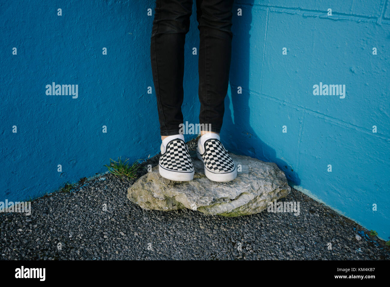 Teenage girl wearing black trousers and checkered canvas shoes, standing on rock against blue wall. Stock Photo