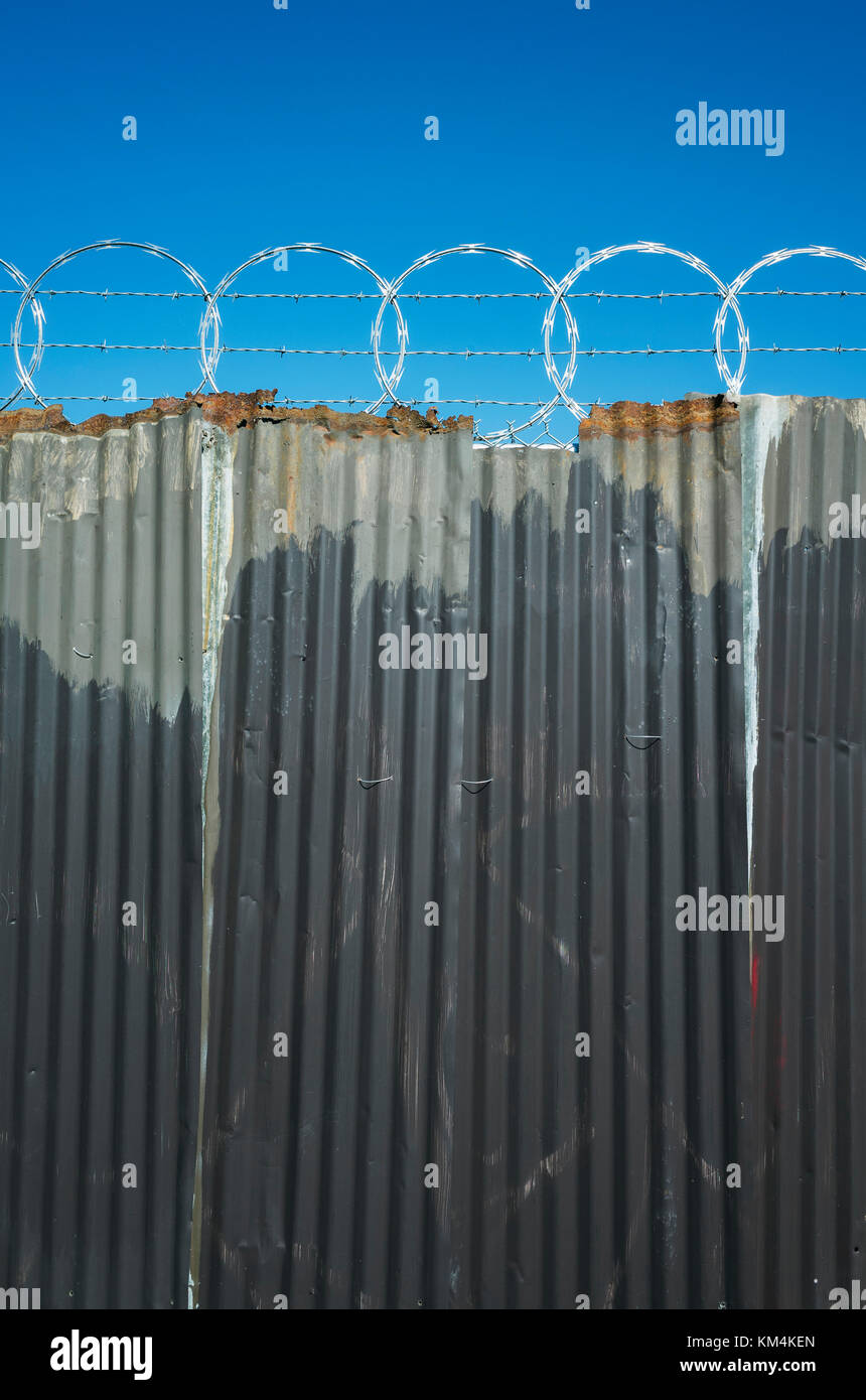 Worn corrugated metal fence, razor wire above. Stock Photo