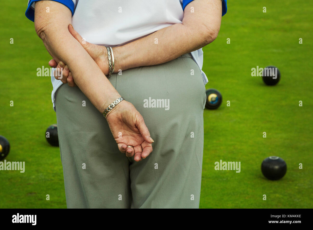 Rear view of a woman lawn bowls player with hands behind her back, and lawn bowls ranged across the playing surface. Stock Photo