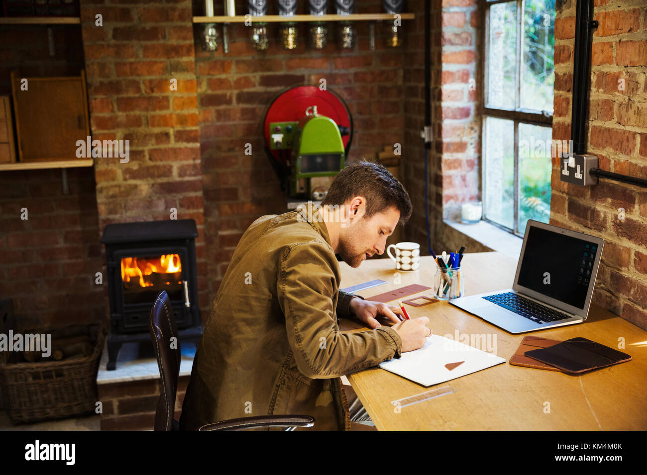 A designer seated in his leatherwork workshop, at a desk drawing on paper. A woodburning stove with a lit fire. Stock Photo