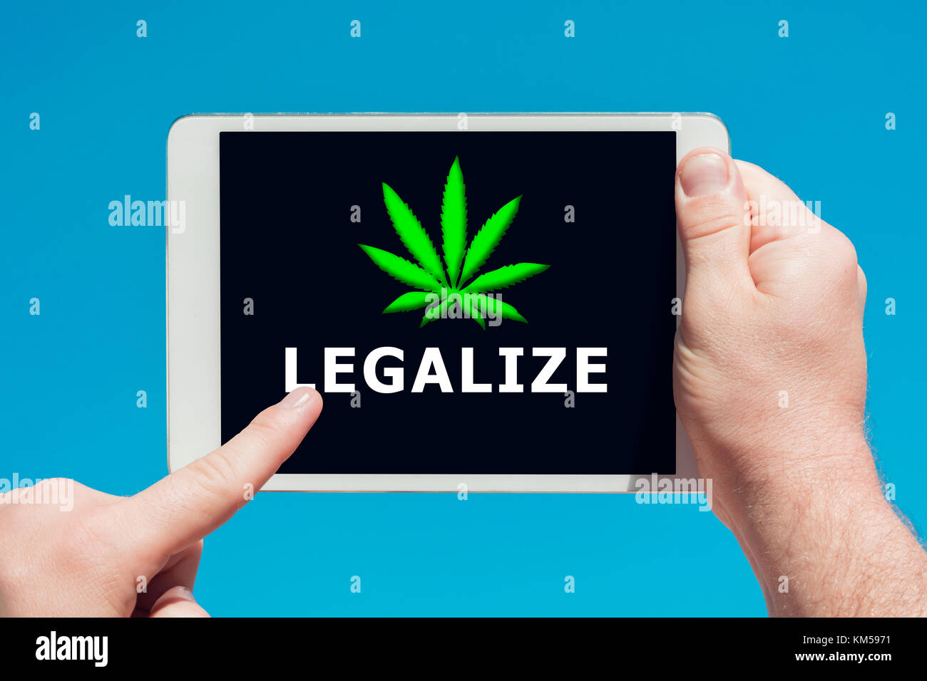 Man holding a tablet device showing a 'legalize marijuana' concept and touching the screen with a finger with blue sky in background. Stock Photo
