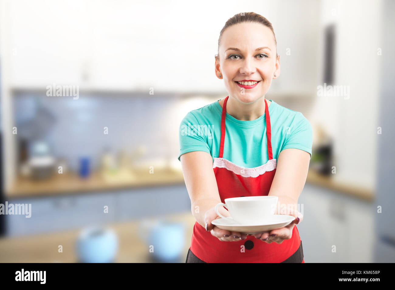 Housemaid or wife offering a cup of coffee or tea on kitchen background wearing red apron Stock Photo