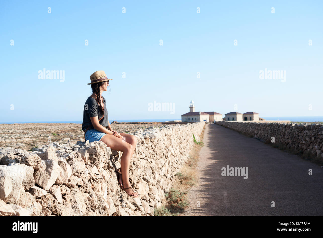 Menorca, Spain, solo traveler sitting in a wall wearing an explorer hat in an empty road to a lighthouse in Menorca island, solo traveler Stock Photo