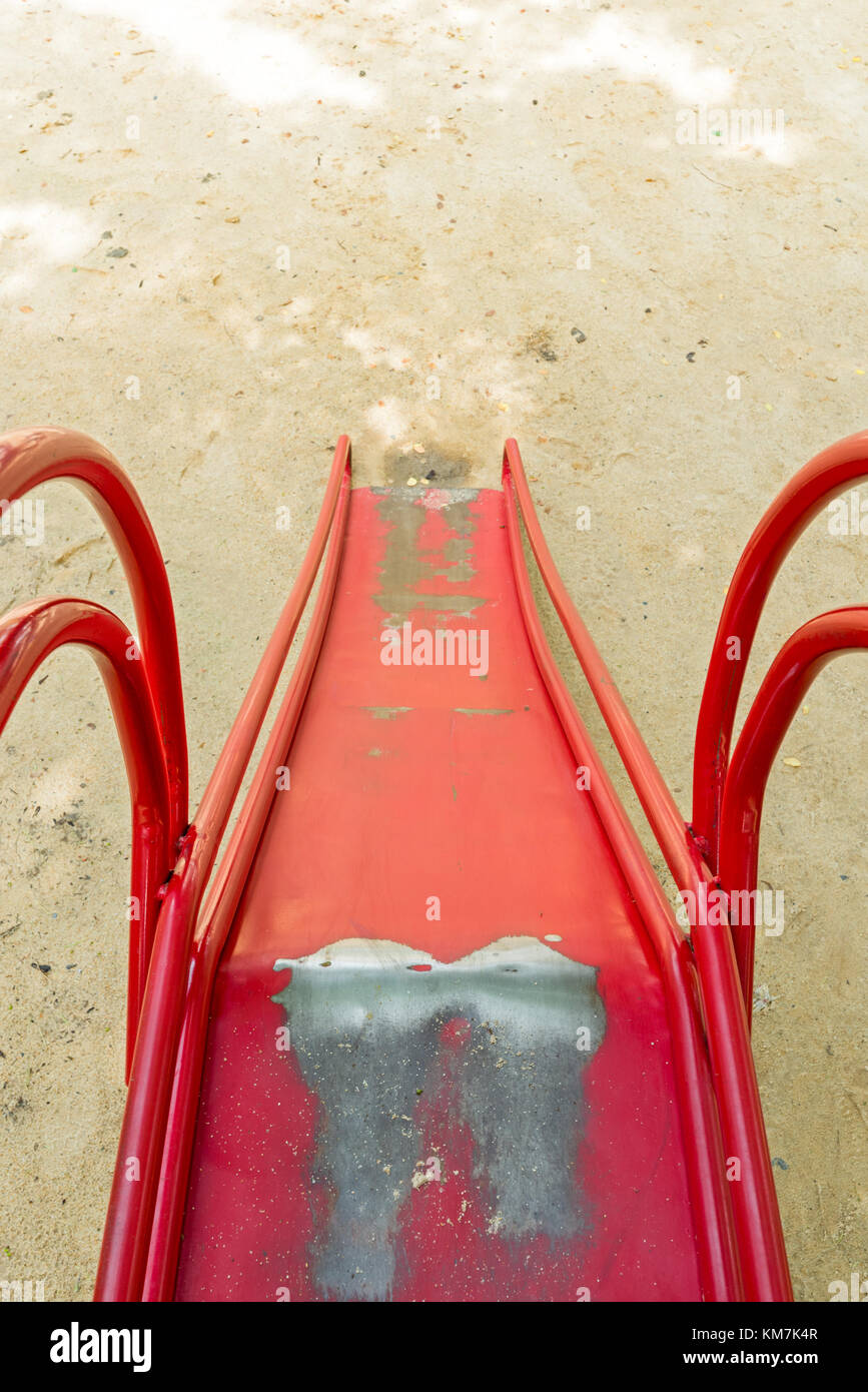 A worn red painted metal child's slide surrounded by a sandy area. Stock Photo