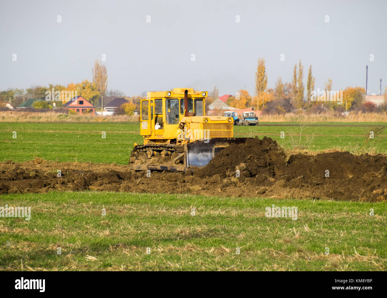 The yellow tractor with attached grederom makes ground leveling. Work on the drainage system in the field. Stock Photo