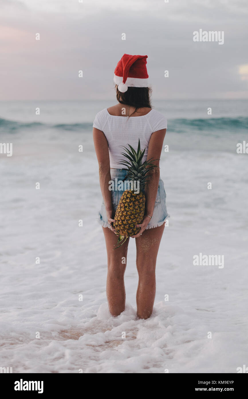 Woman wearing a Christmas Santa hat standing in ocean surf holding a pineapple behind her back, Haleiwa, Hawaii, United States Stock Photo