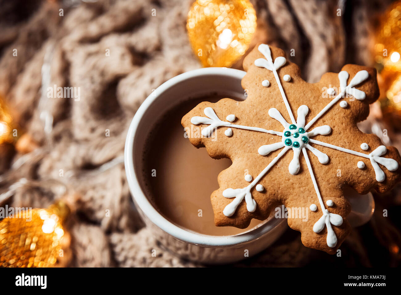 Christmas background with coffee in a white cup and gingerbread snowflakes on a brown knitted winter scarf and a glowing golden garland. Beautiful con Stock Photo