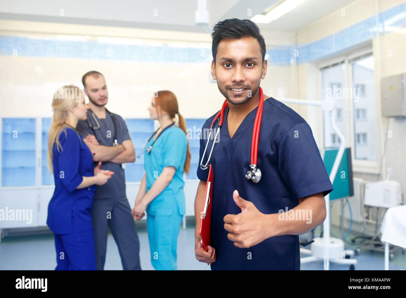 Asian male doctor in front of team, looking at camera, show thumb up, with medical team in background. Multiracial team of young doctors in a hospital standing in a operating room. Stock Photo