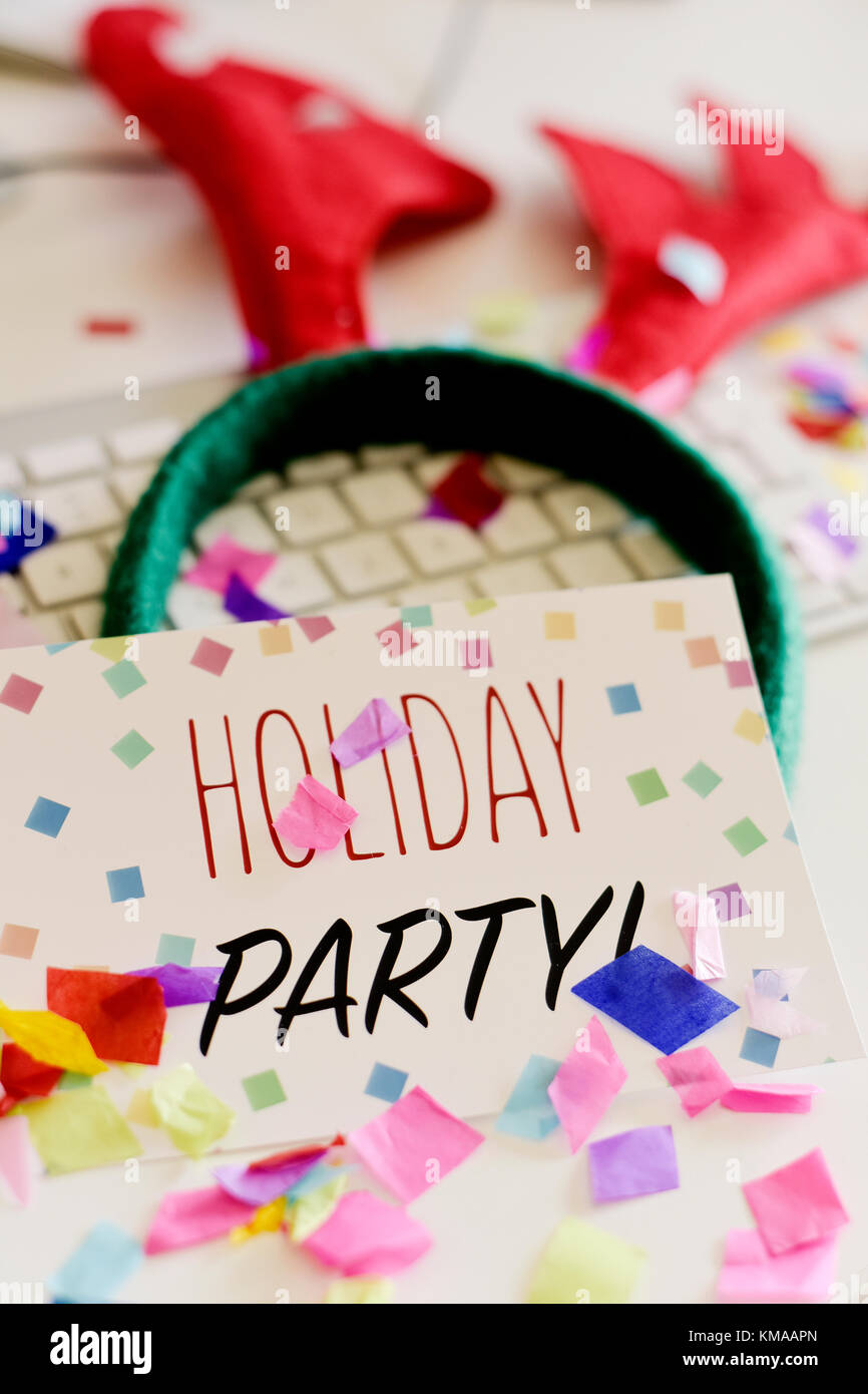 a signboard with the text holiday party, on an office desk full of confetti, next to a reindeer antlers headband and a computer keyboard Stock Photo