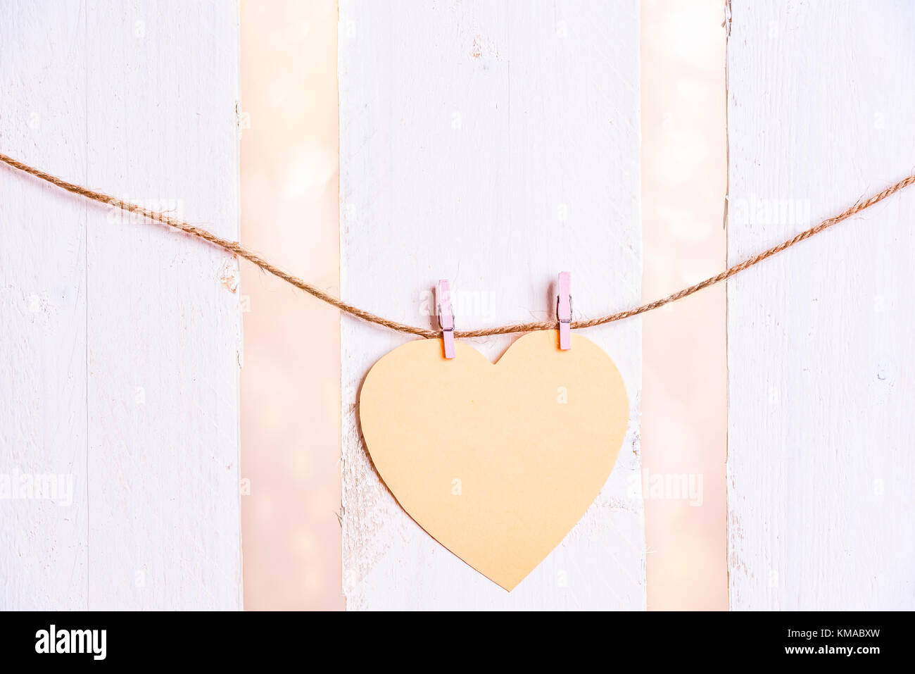 Romantic image with a yellow heart-shaped paper note tied to a string with two wooden clips, on a white wooden fence background. Stock Photo