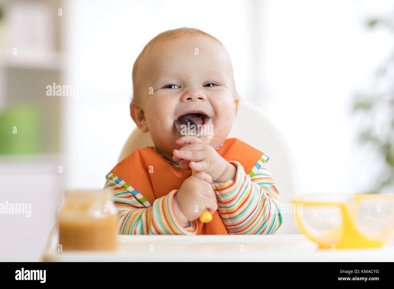 Cheerful baby child eats food itself with spoon. Portrait of happy kid boy in high-chair. Stock Photo