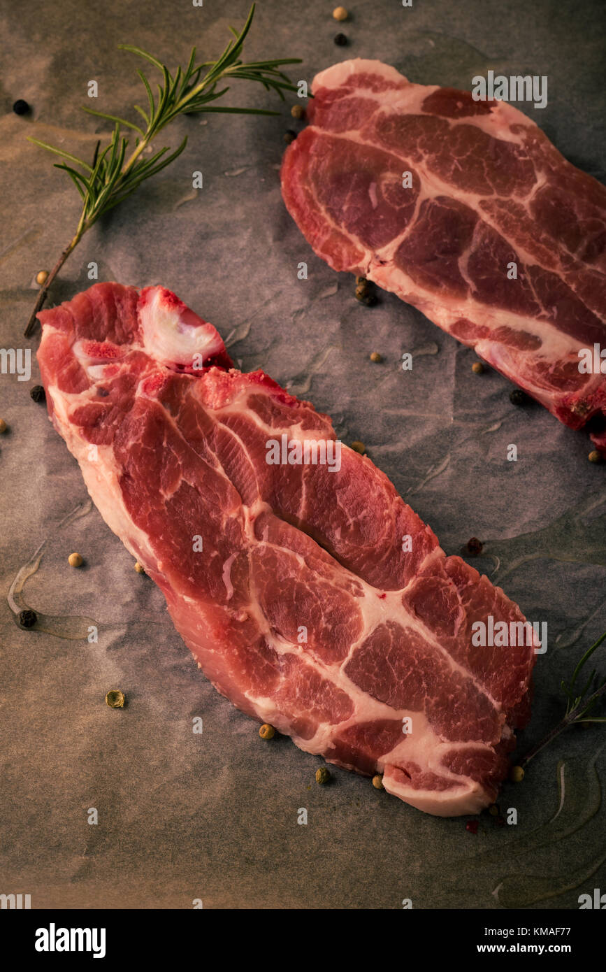 Vertical photo of two slices of pork neck steaks. The meat portions with red color and fat is placed on worn baking sheet of paper with pepper, rosema Stock Photo