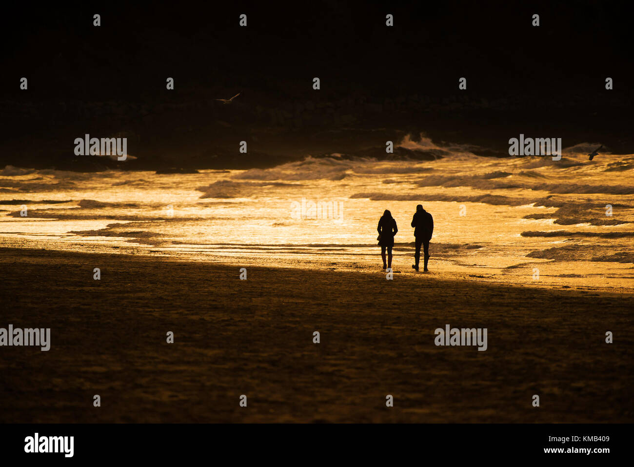 Sunset - a couple seen in silhouette walking along the shore line during an intense sunset at Fistral Beach Newquay Cornwall UK. Stock Photo