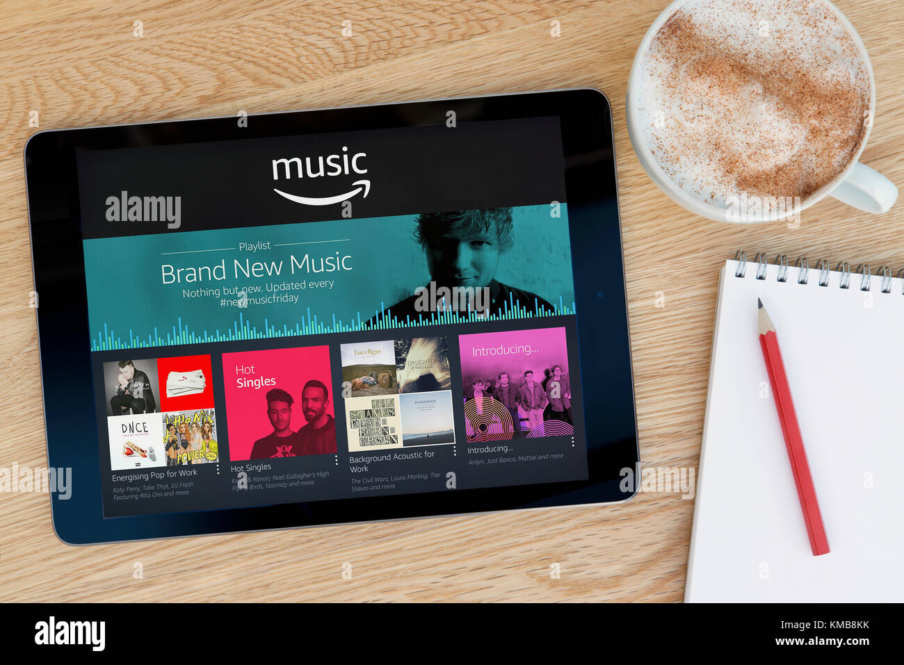 The Amazon Music app features on an iPad tablet device which rests on a wooden table beside a notepad and pencil and a cup of coffee (Editorial only). Stock Photo