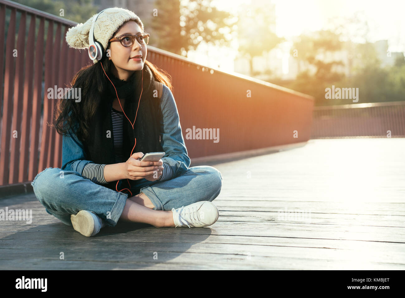 stylish charming asia woman enjoys smartphone music on the city bridge, girl enjoys sunny day in the winter season. Stock Photo