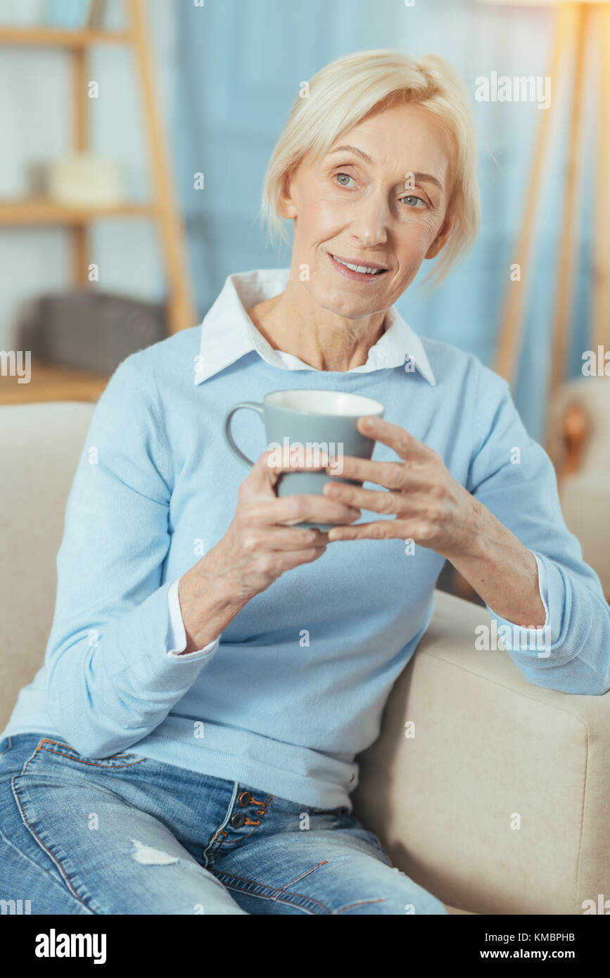 Thoughtful senior woman sitting on a sofa and drinking hot tasty tea Stock Photo