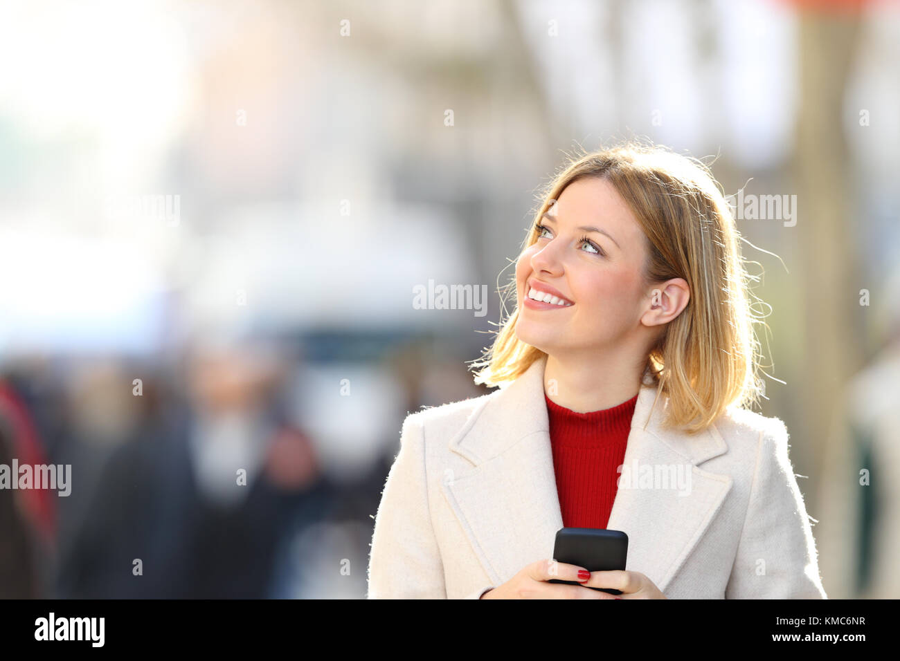 Portrait of a happy woman holding a smart phone thinking and looking at side on the street in winter Stock Photo